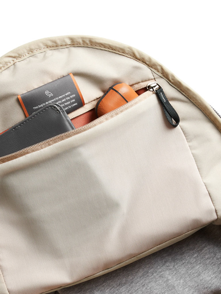 A close-up shows the neatly organized inner pocket of the Bellroy Classic Backpack Plus - 24L, made from water-resistant recycled fabric, holding a gray wallet, a brown cylindrical case, and a visible fabric tag with text.