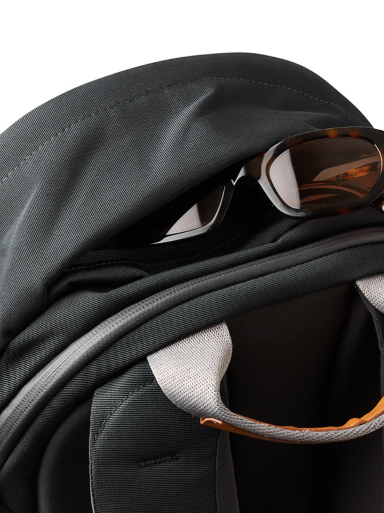 A close-up of the Bellroy Classic Backpack Plus - 24L, made from water-resistant recycled fabric in black, with brown sunglasses in the front zipper pocket. It features padded straps—one with a light gray accent—set against a white background.