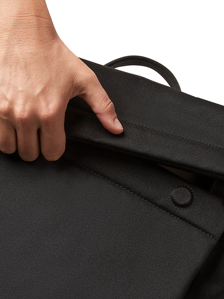 A close-up of a hand lifting the flap of the Bellroy Melbourne Backpack Compact 13L in black, featuring a button closure, set against a white background.