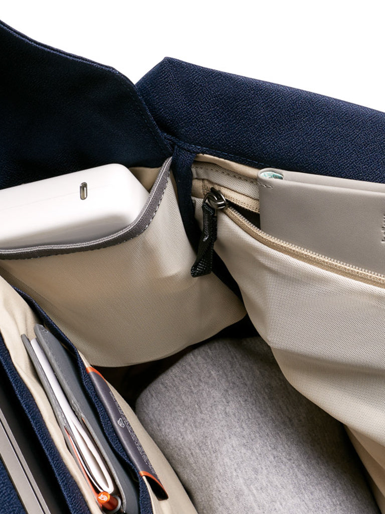 A close-up of the organized interior of the Bellroy Melbourne Backpack Compact 13L shows multiple beige pockets holding a charger, notebook, and pens, all set against a dark blue fabric lining.