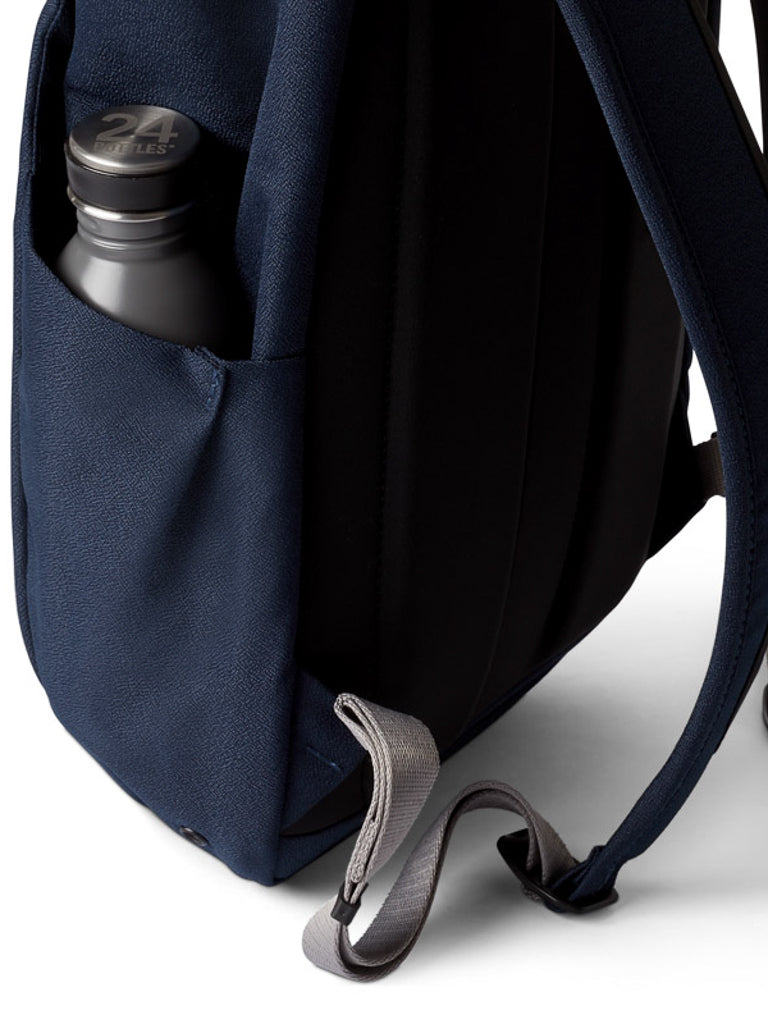 A close-up of the Bellroy Melbourne Backpack Compact 13L in navy blue highlights its side pocket with a black reusable water bottle, padded black straps, a gray adjustment strap, and minimalist design on a white background.