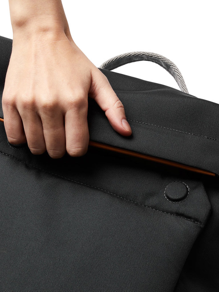 A hand lifts the flap of the Bellroy Melbourne Backpack Compact 13L, revealing the edge of a slim orange electronic device inside. The black bag features a fabric handle and a snap button closure.