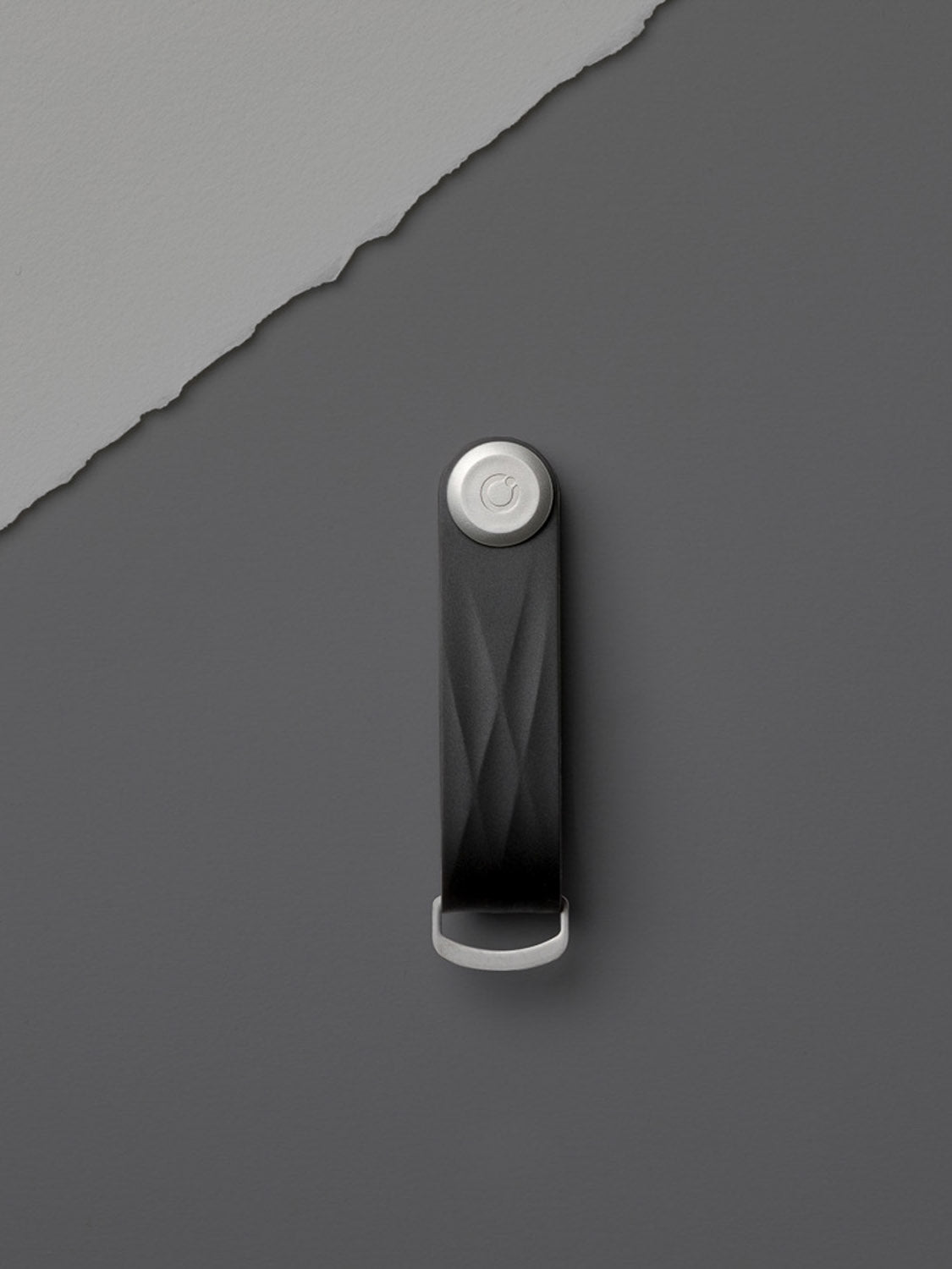 The Orbitkeys Key Organizer Rubber Active, in black and silver with a textured finish, rests on a gray surface, highlighted by a torn light gray paper edge in the top left.