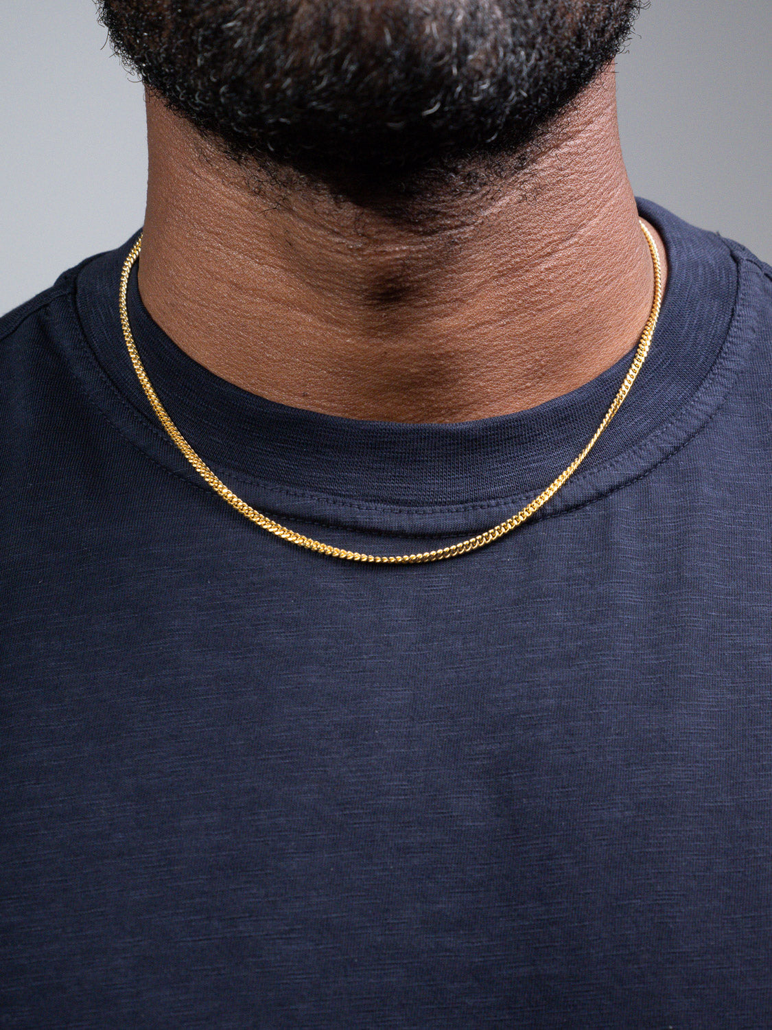 A close-up of a person wearing a navy blue shirt and the Blaze 100 Miami Cuban Necklace by ID Menswear, focusing on the neck and upper chest with a neatly groomed beard visible at the top.