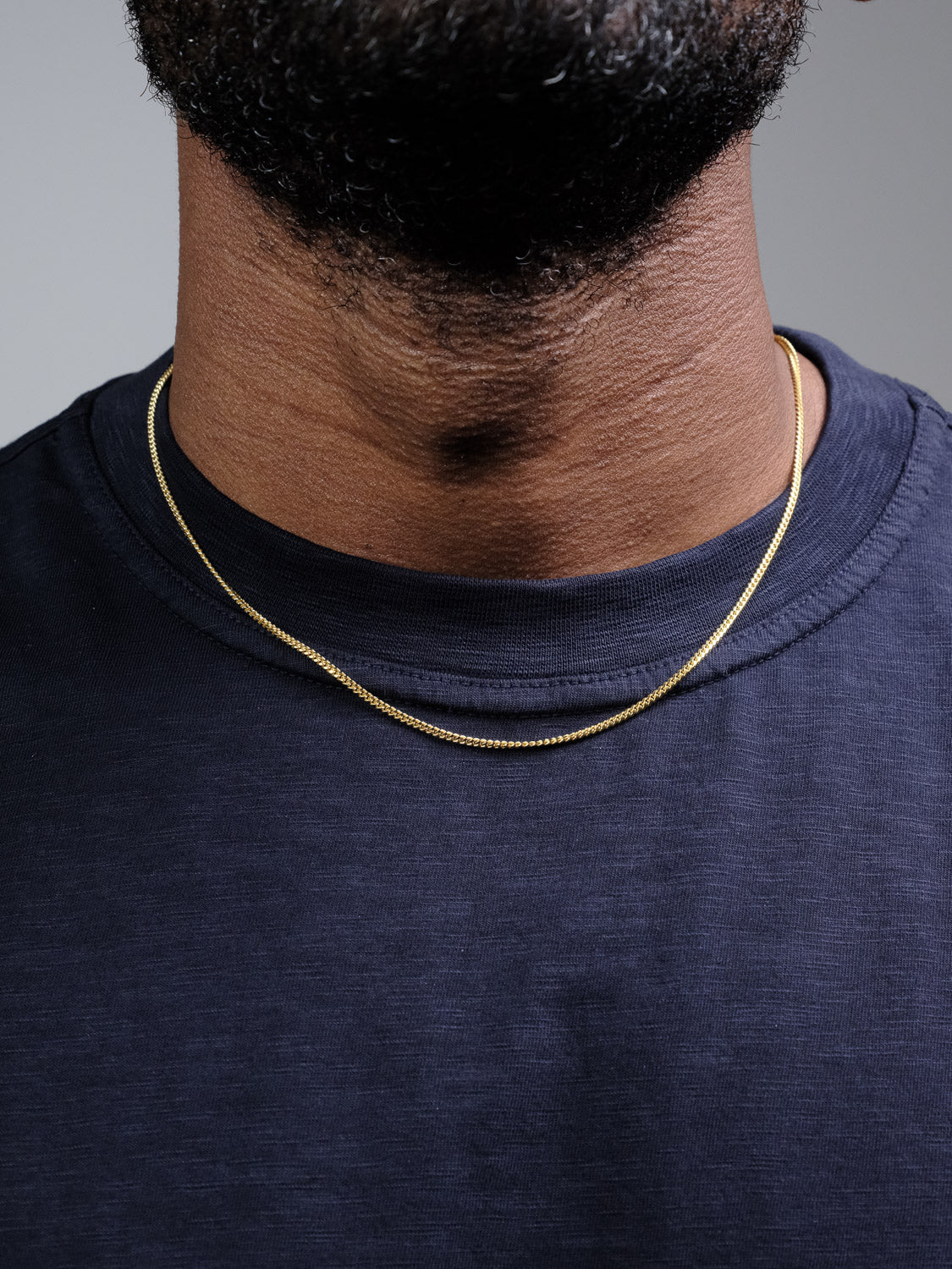 A close-up of a person’s neck and jaw wearing the Blaze 60 Miami Cuban Necklace by ID Menswear with a thin gold-plated chain and a dark navy T-shirt, set against a plain, neutral background.