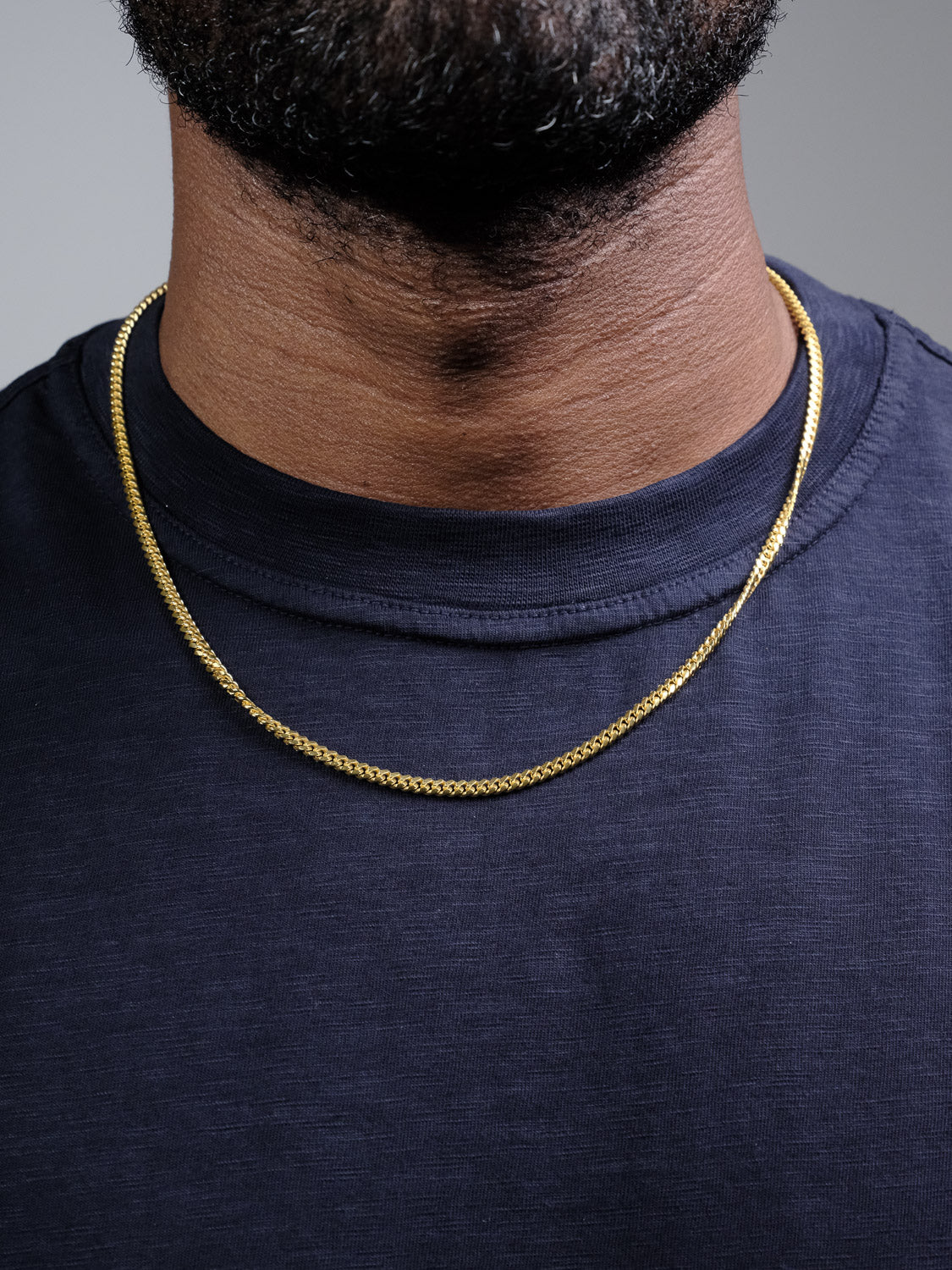 A close-up of a person's neck wearing the Blaze 80 Miami Cuban Necklace by ID Menswear with a dark blue crew neck t-shirt. The background is plain and neutral.
