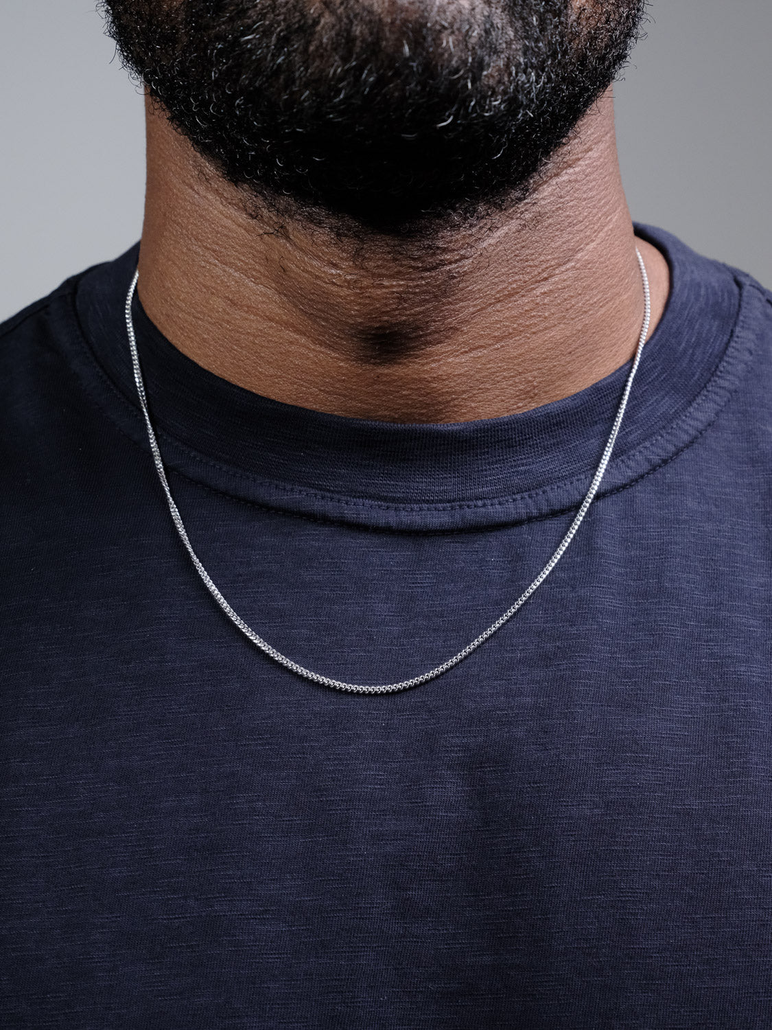 Close-up of a person wearing a dark t-shirt and the ID Menswear Blaze 60 Miami Cuban Necklace in sterling silver. Only the lower face, neck, and upper chest are visible against a neutral background.
