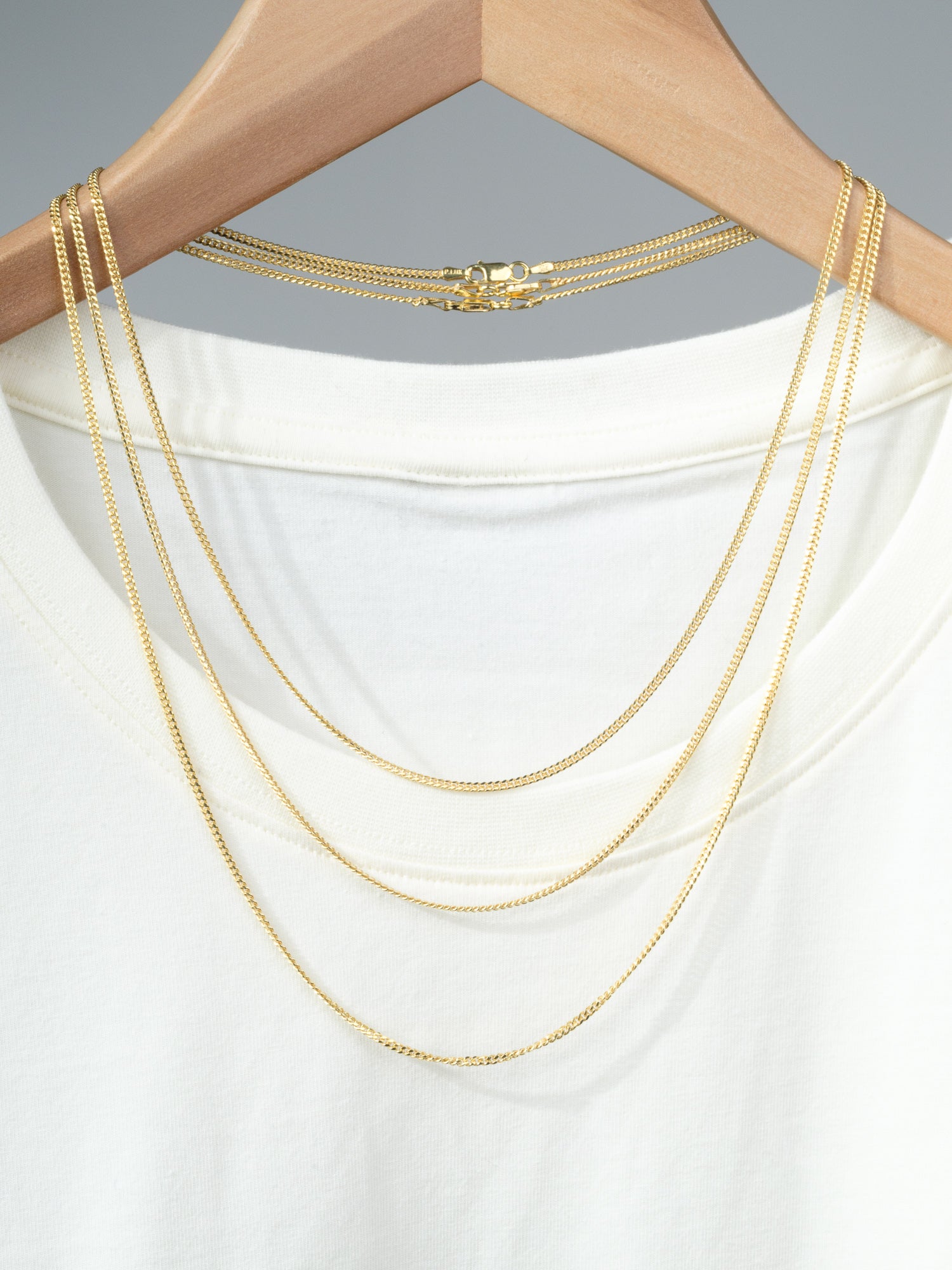 A white T-shirt on a wooden hanger displays three Blaze 60 Miami Cuban Necklaces by ID Menswear, each gold plated and draped over the shoulders and neckline. The background is neutral gray.
