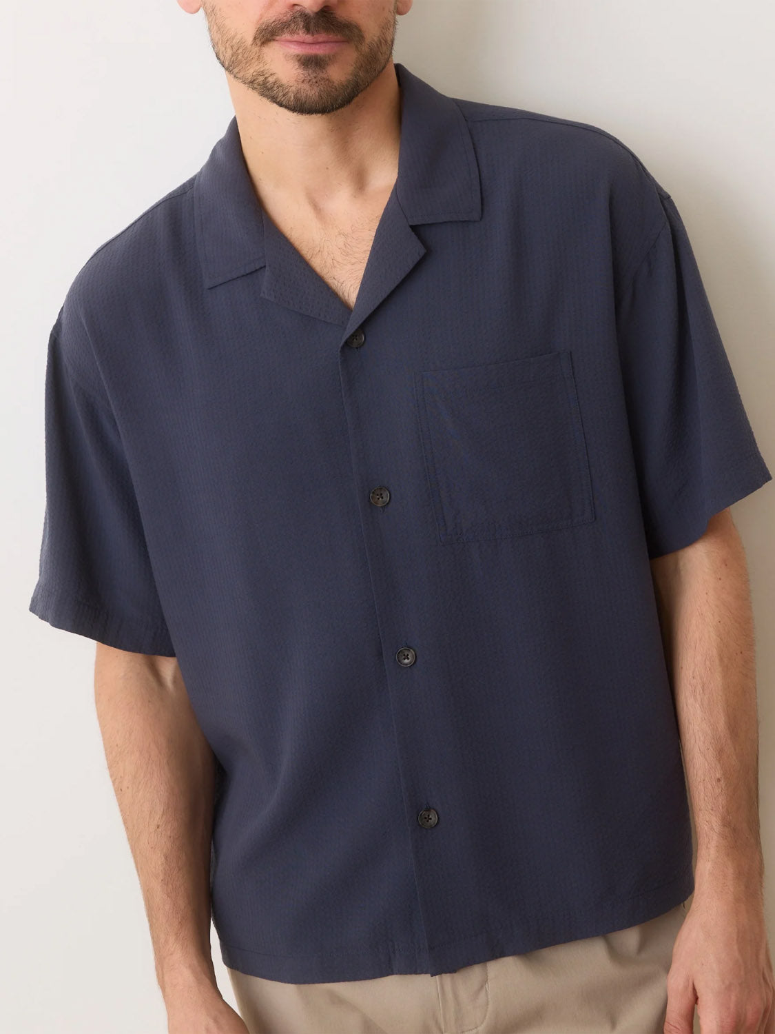 A person in a Marine Layer Boxy Resort Shirt, dark blue with short sleeves, a resort collar, and front pocket stands against a light wall. The light-colored pants complement the TENCEL Lyocell fabrics smooth texture, with only the upper body visible.
