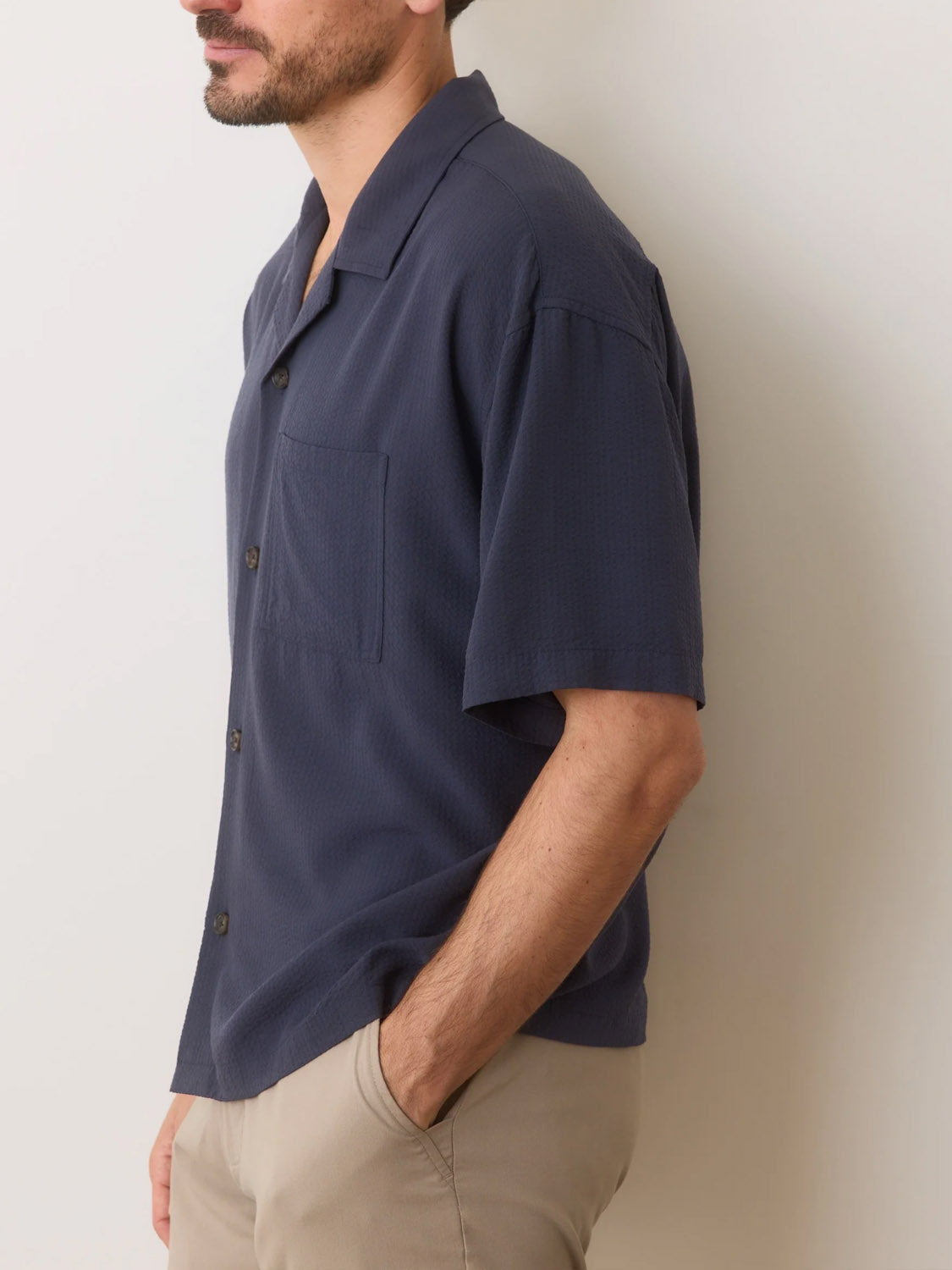 A man wearing a Marine Layer Boxy Resort Shirt, a dark TENCEL Lyocell short-sleeve button-up with a pocket, stands in profile with one hand in the pocket of his light pants against a plain background.