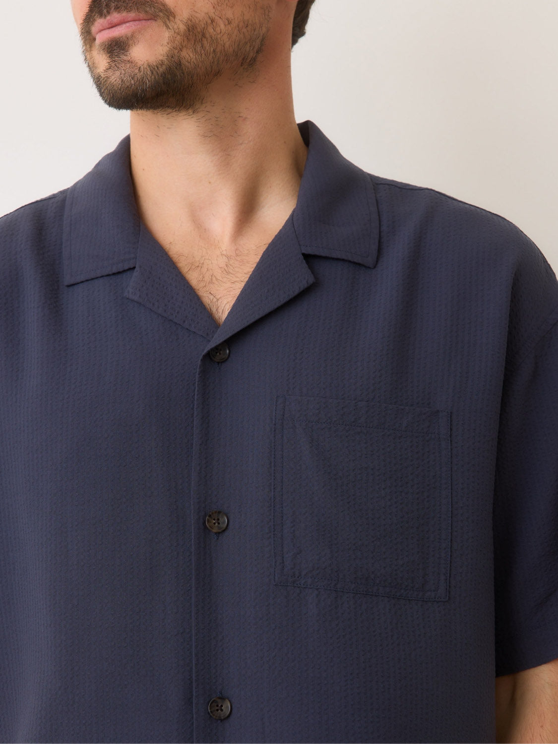 A person in a Marine Layer Boxy Resort Shirt stands against a plain background. The dark blue, short-sleeve, button-down shirt by Marine Layer features a textured finish and chest pocket. Their face is partially visible with facial hair.