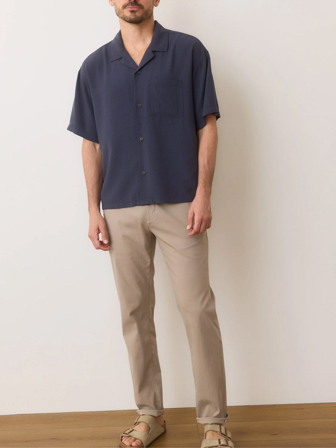 A person in a Marine Layer Boxy Resort Shirt in navy, paired with beige pants and sandals, stands on a wooden floor against a light-colored wall.