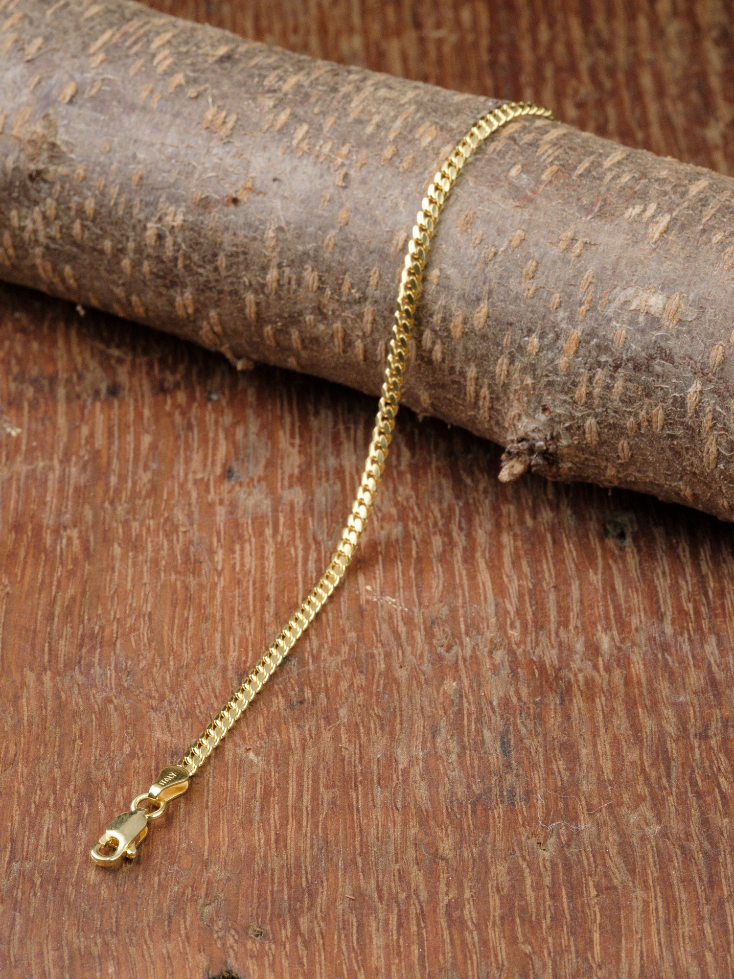 The Blaze 80 Miami Cuban Bracelet by ID Menswear, featuring a thin gold-plated chain and lobster clasp, is displayed draped over a textured log on a rustic wooden surface.