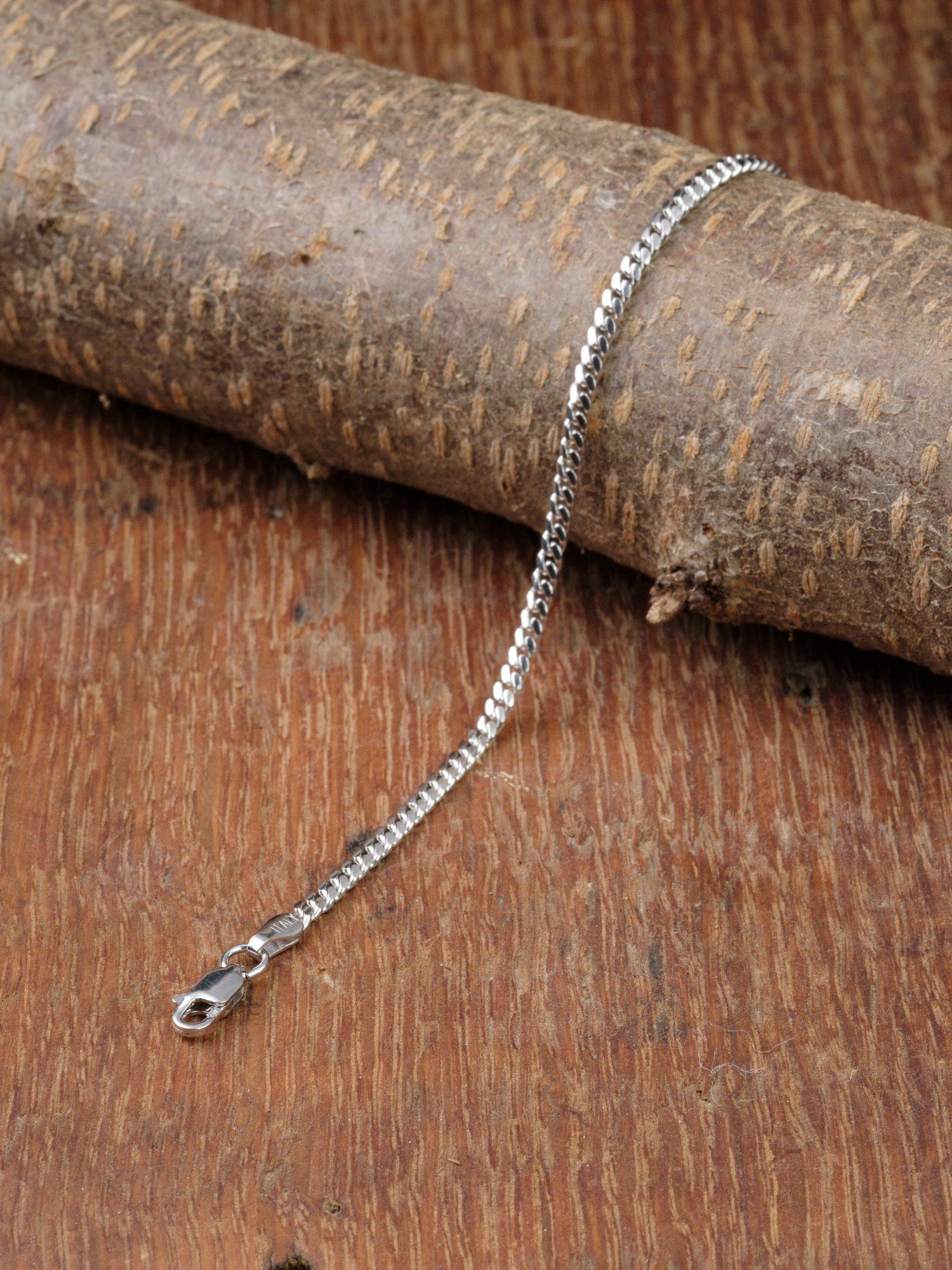 The Blaze 80 Miami Cuban Bracelet by ID Menswear, a sleek silver chain with a lobster clasp, is showcased draped elegantly over a textured wooden surface.