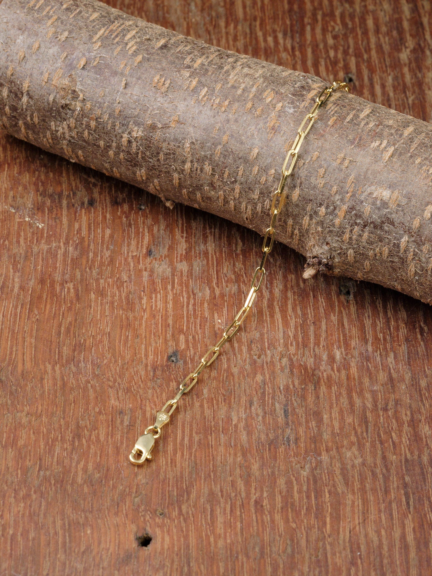 The Kai 80 Bracelet by ID Menswear, featuring an elongated gold-plated paperclip chain design, is displayed on textured wood set against a wooden background.