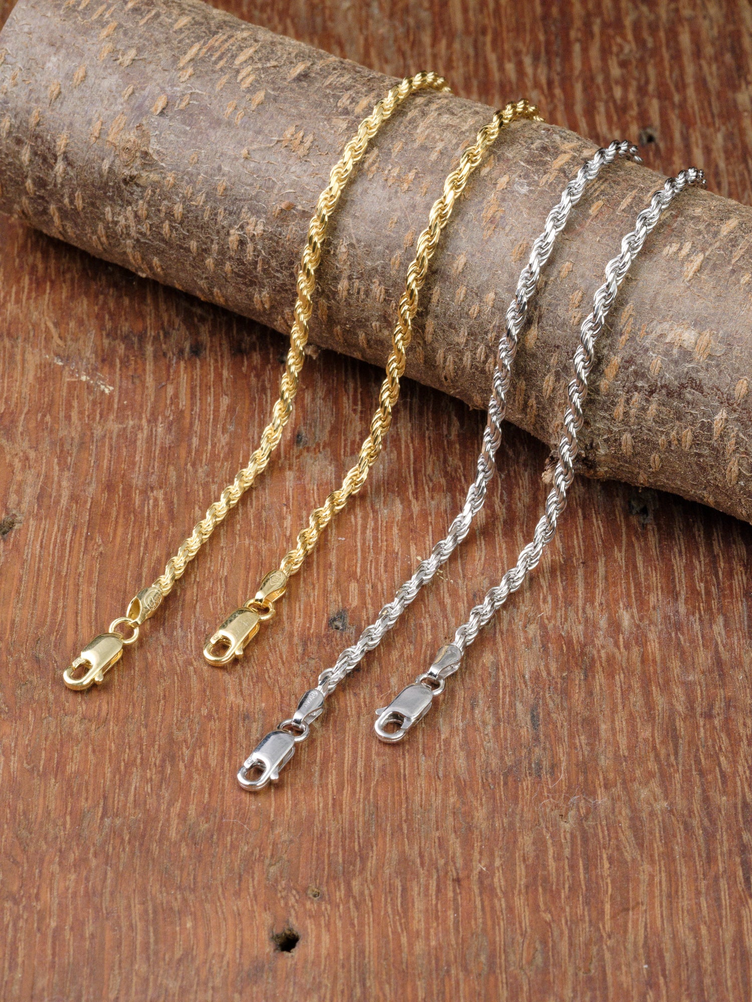 Two Orion 50 Bracelets by ID Menswear, featuring gold and 925 sterling silver rope chains with clasps, are elegantly draped over a cylindrical piece of wood on a textured wooden surface.