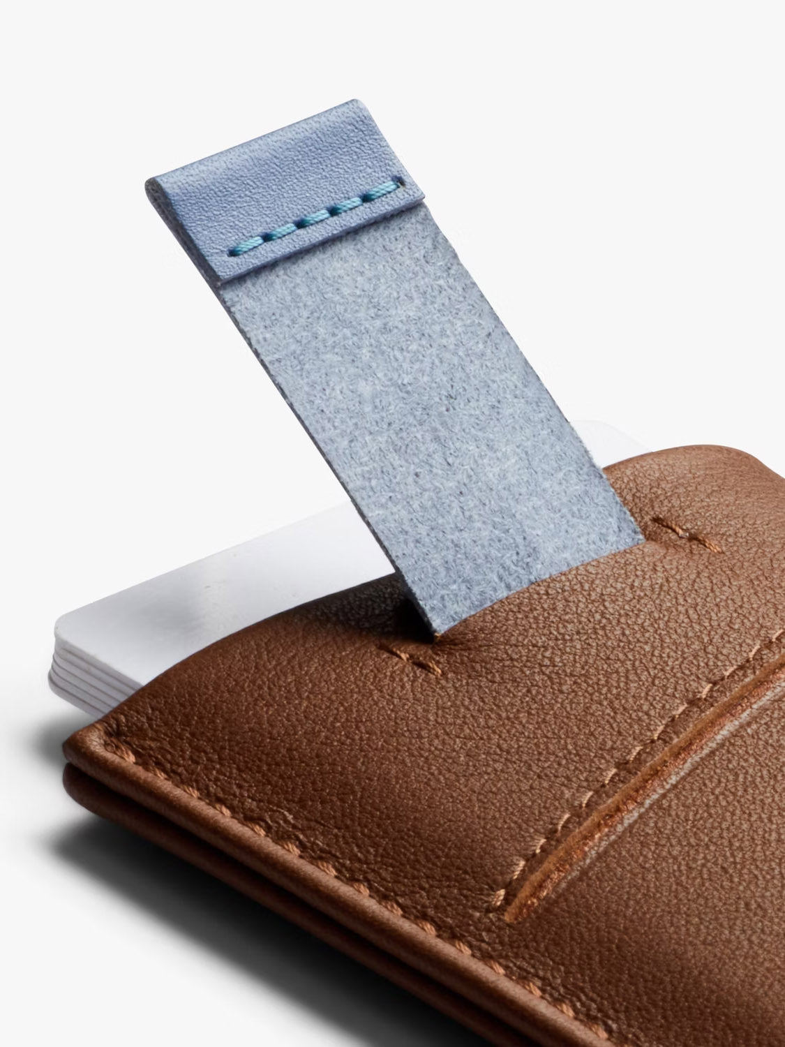 A close-up of the Bellroy Card Sleeve Wallet in brown leather shows its textured blue pull tab partially raised, revealing a stack of white cards inside the main pocket.