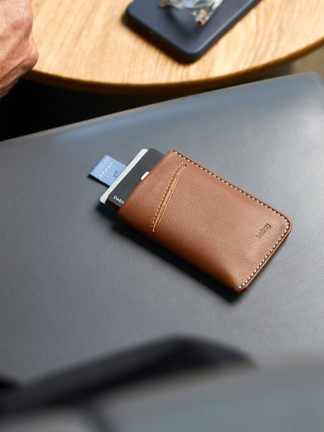 A Bellroy Card Sleeve Wallet in brown, with three cards partially visible, rests on a gray surface. A wooden table and a hand are blurred in the background.
