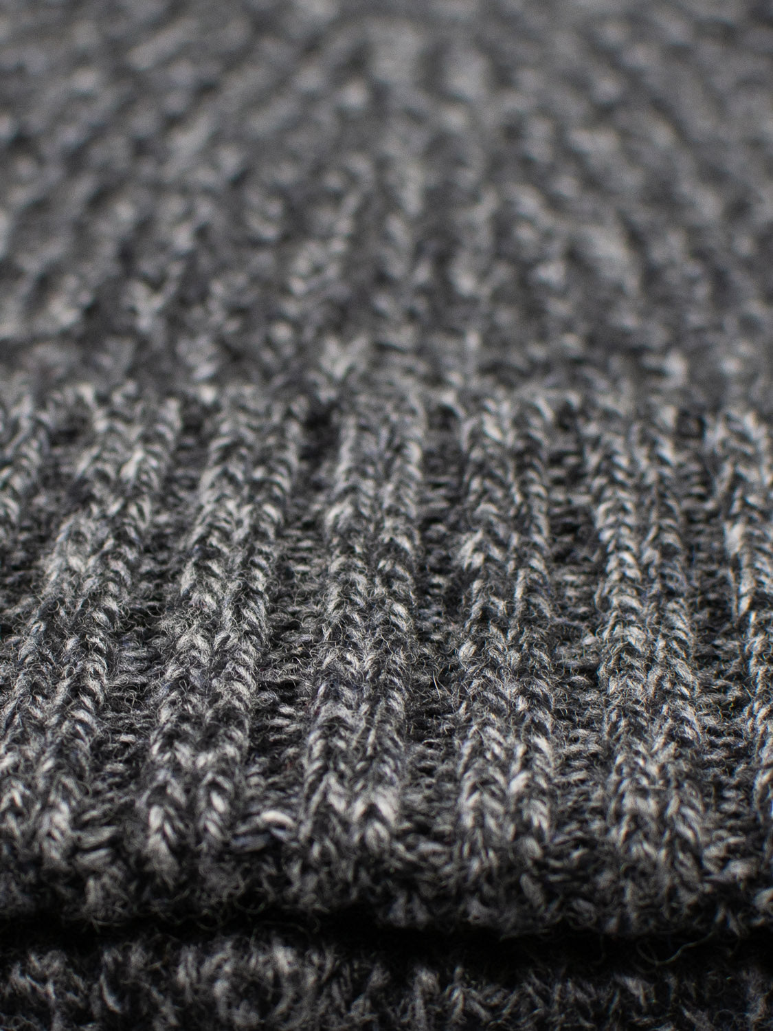 A close-up of the Cho Oyu Merino Wool Fishermans Beanie by ID Menswear reveals a ribbed, textured grey knit. The thick yarn blends light and dark grey shades, offering a cozy look ideal for winter.
