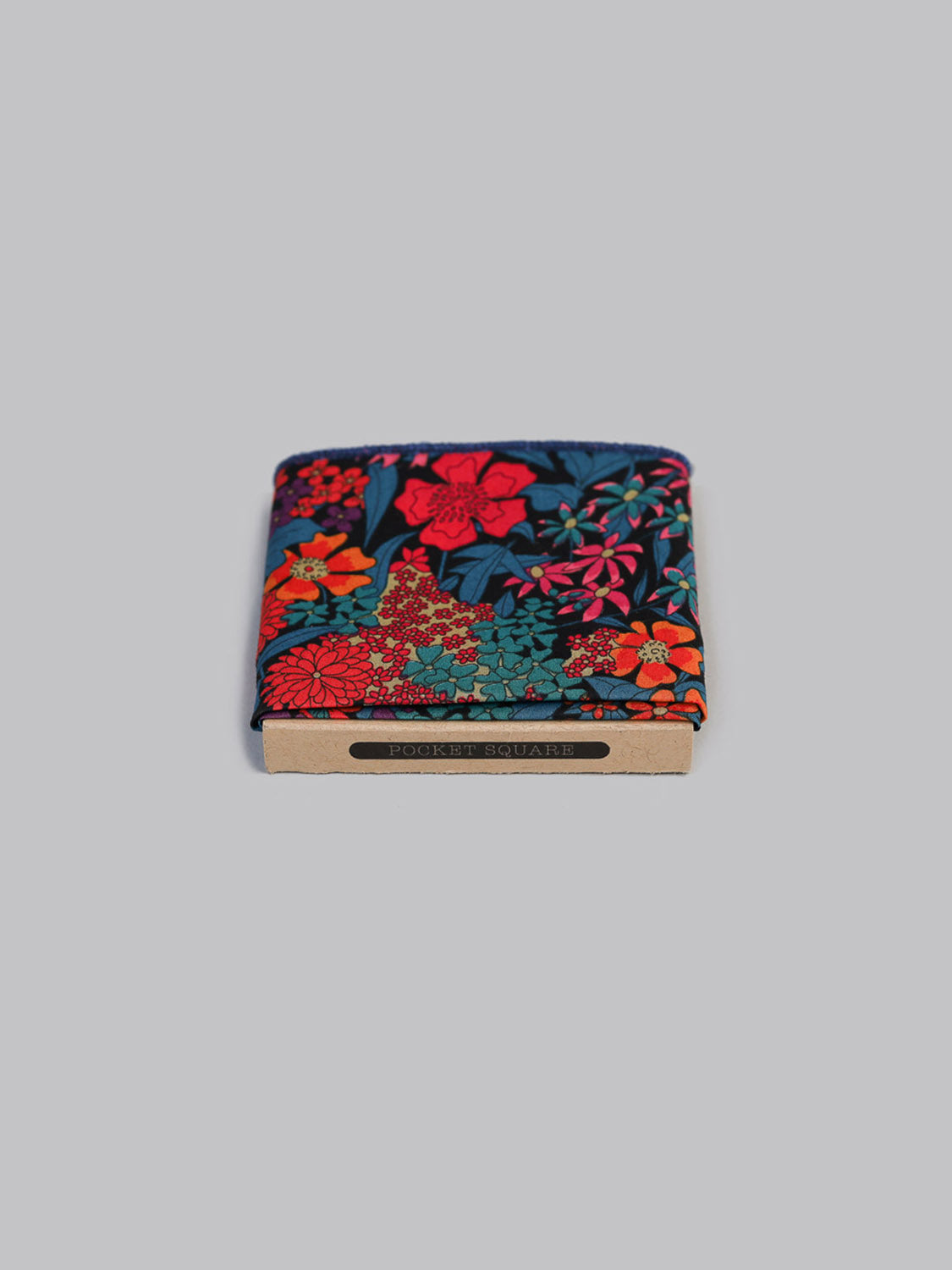 A folded ID Menswear Liberty™ Ciara Pocket Square in vibrant red, pink, and teal floral Tana Lawn cotton rests in a brown holder labeled POCKET SQUARE, set against a light gray background.