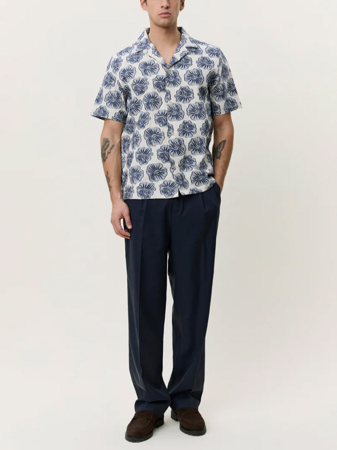 A man faces forward against a light background, wearing the Les Deux Dahlia Jacquard SS Shirt in blue pattern, paired with dark blue loose-fit pants and dark shoes.