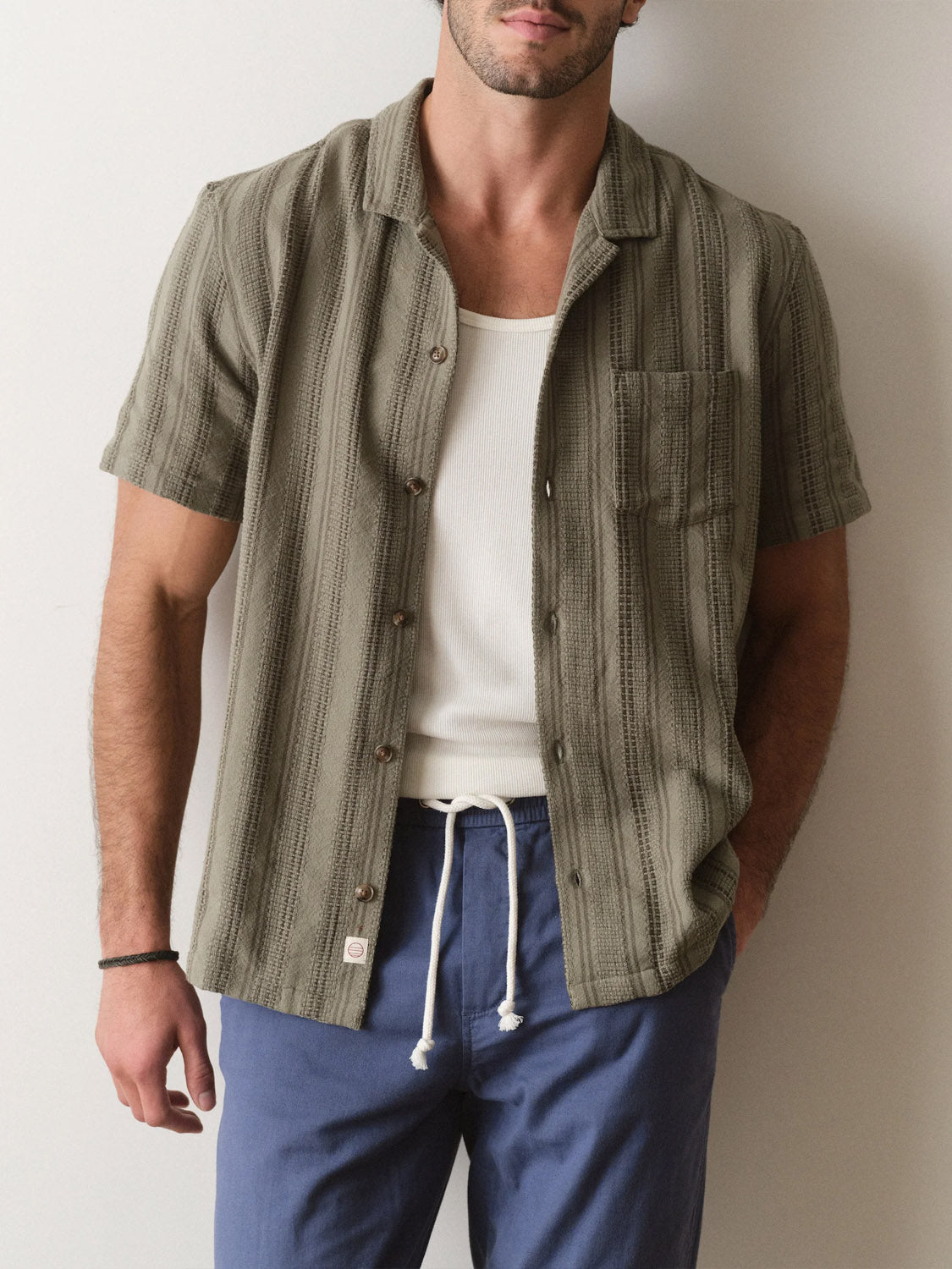 A man stands against a plain, light wall, wearing Marine Layer’s Dobby Resort Shirt in olive green over a white tank and blue drawstring pants, with one hand in his pocket.