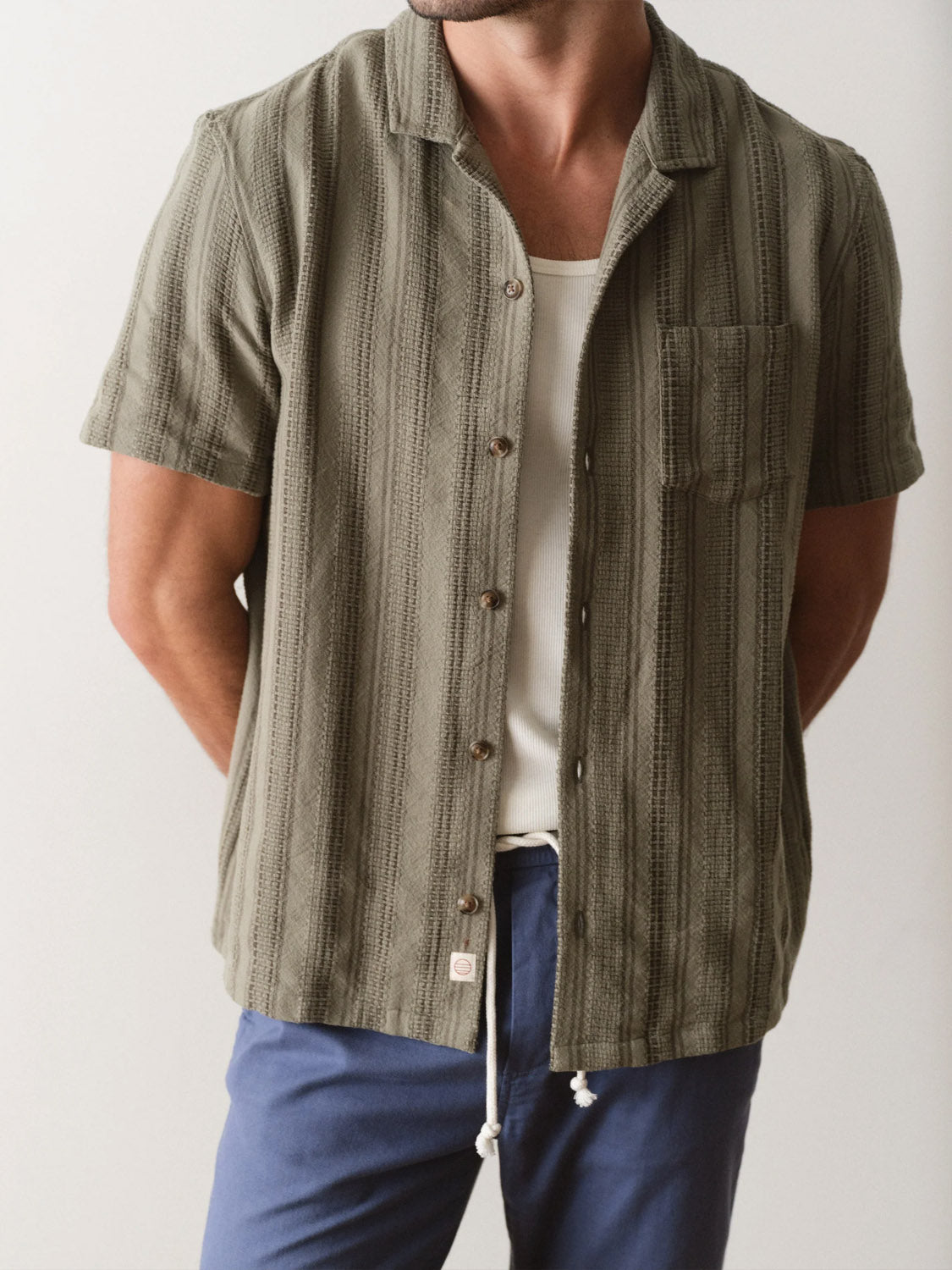 A man stands against a plain background with his arms behind his back, wearing Marine Layer’s green Dobby Resort Shirt—short-sleeved with a subtle stripe—over a white undershirt and blue drawstring pants.