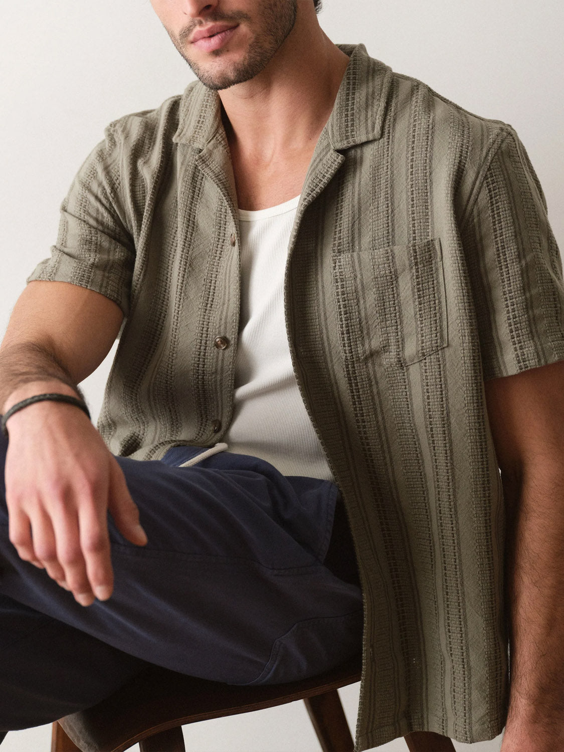 A man sits casually with one arm on his knee, wearing Marine Layer’s Dobby Resort Shirt in olive green over a white tank and navy blue pants. His face is partly out of frame as he relaxes in the chair.