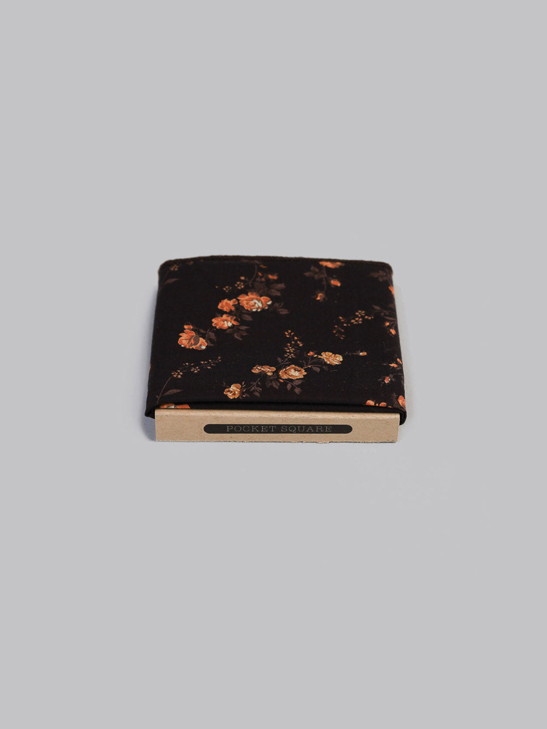 The Liberty™ Elizabeth Pocket Square by ID Menswear, crafted from Liberty of London Tana Lawn cotton in an orange floral pattern, is folded and presented in a cardboard holder labeled Pocket Square on a light gray background.