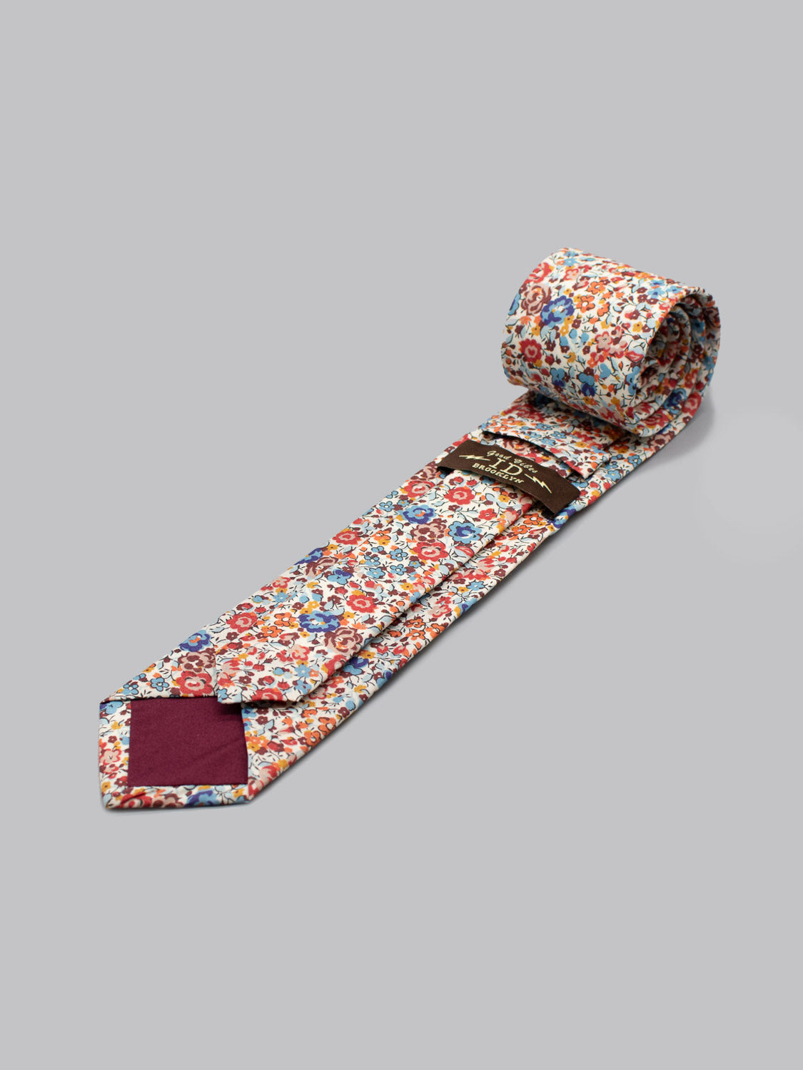 A Liberty of London Tana Lawn™ Emma Rouge tie by ID Menswear, handmade in Brooklyn from soft cotton with a vibrant red, blue, and yellow floral pattern on white, is rolled up on a light gray surface with its pointed end facing forward.