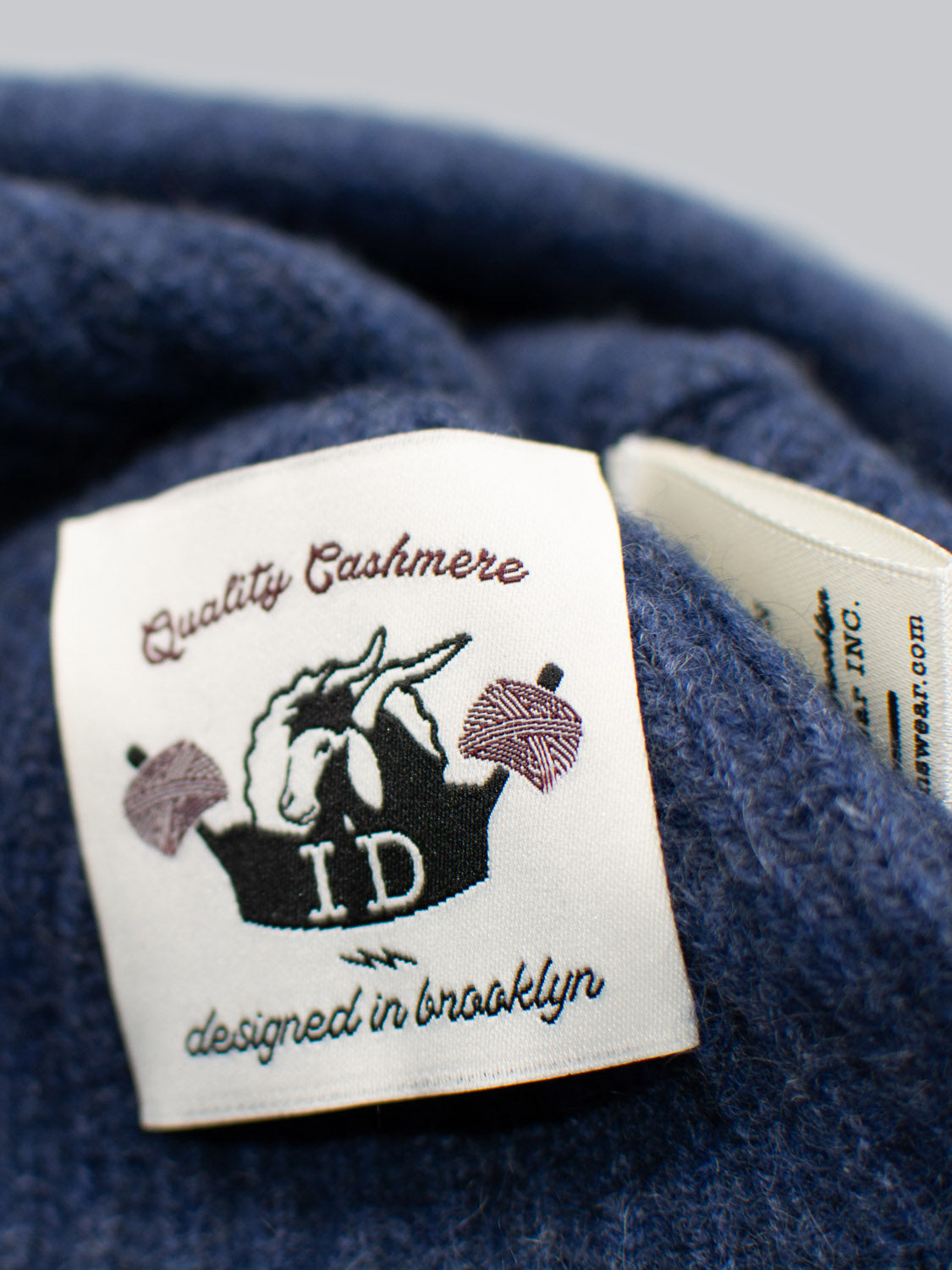 A close-up of the Everest Cashmere Beanie by ID Menswear shows a label on blue fabric with a unicorn and yarn balls, reading “Quality Cashmere I.D. designed in brooklyn”—blending style and lightweight comfort.