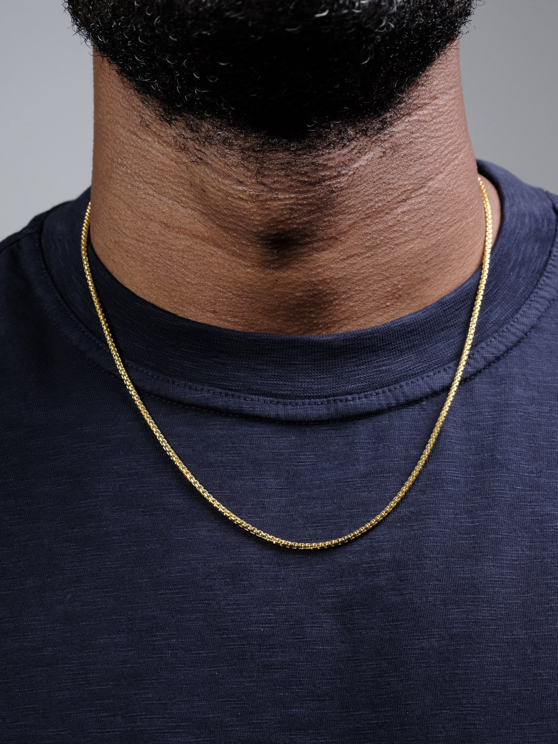 A close-up shows a person's neck in a navy blue shirt, wearing the delicate Ezra 40 Box Chain Necklace by ID Menswear. The lower face and beard are partially visible, highlighting the thin gold-plated design.