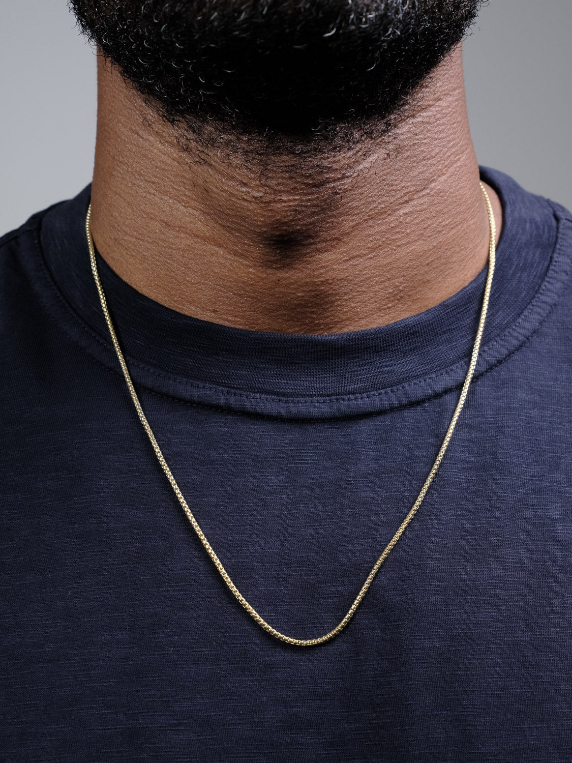 A close-up of a person wearing the Ezra 35 Box Chain Necklace by ID Menswear and a dark blue t-shirt, with the lower part of their bearded face visible.