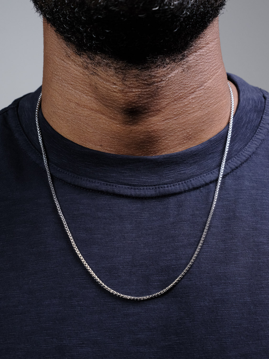 A close-up shows a person wearing a navy blue shirt and the Ezra 40 Box Chain Necklace by ID Menswear (925 Sterling Silver) around their neck. Only the lower part of the face is visible; no additional details are shown.
