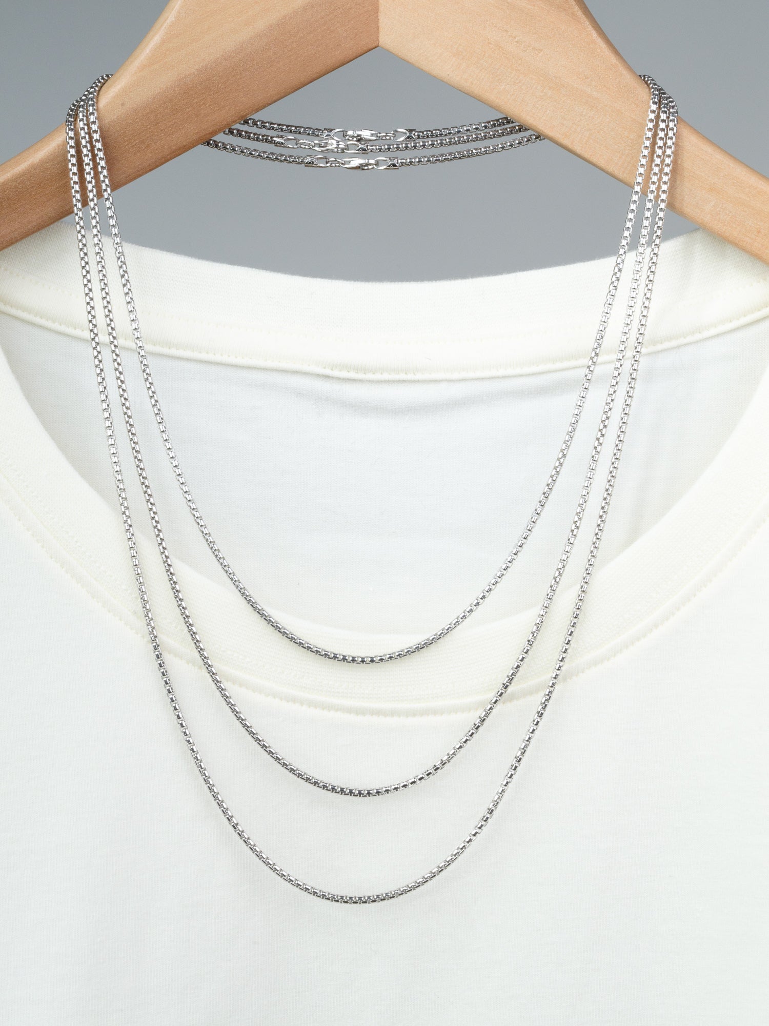 The Ezra 40 Box Chain Necklace by ID Menswear, crafted from 925 Sterling Silver in Italy, is showcased layered on a white T-shirt hung on wood, set against a neutral gray background.