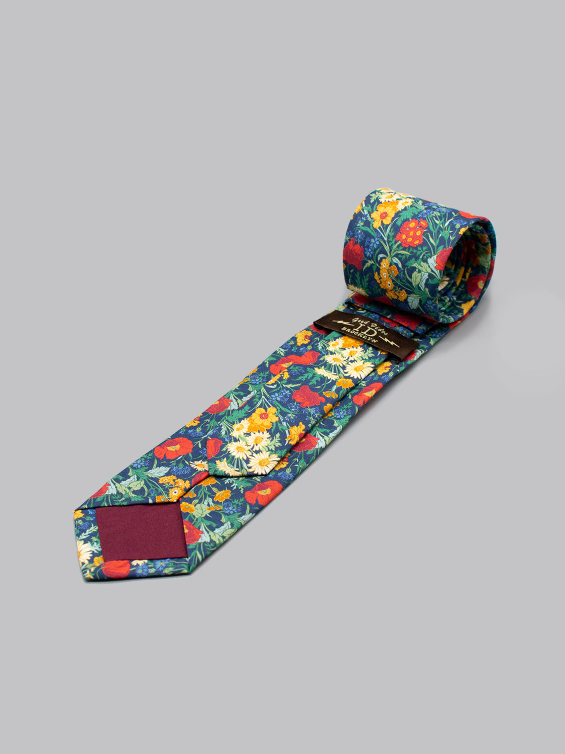 The ID Menswear Liberty of London Tana Lawn™ Florence tie features a vibrant floral pattern in red, yellow, and green on blue, handmade in Brooklyn with a solid burgundy tip. Photographed on a plain gray surface.