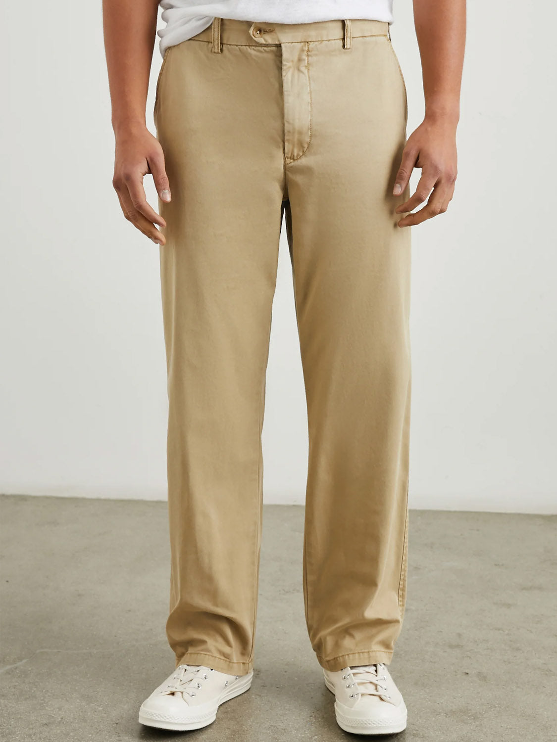 A person is standing in Rails Garrett Pant in True Khaki and white sneakers. Their upper body is out of frame, against a plain light-colored wall and gray floor.