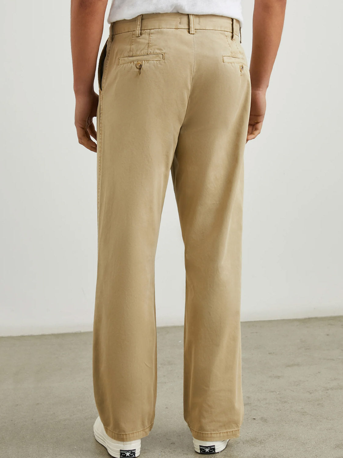 A person stands on a grey floor, seen from behind in Rails Garrett Pant True Khaki cotton chino pants, a white shirt, and white sneakers.