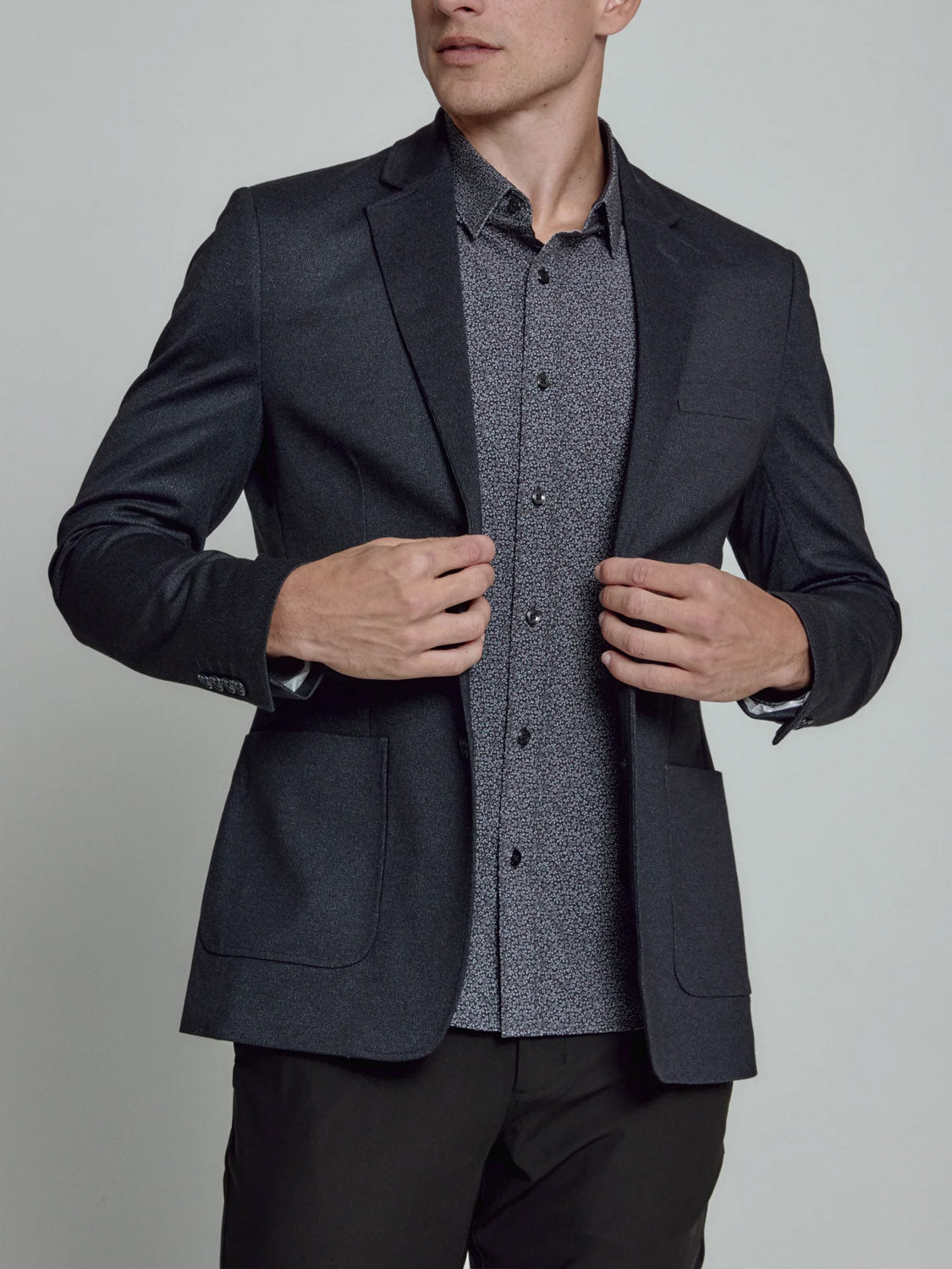 A man adjusts the 7 Diamonds Gatsby Blazer, featuring a notched lapel, over a patterned shirt and dark pants. He stands against a plain light background, with his face partially out of frame.