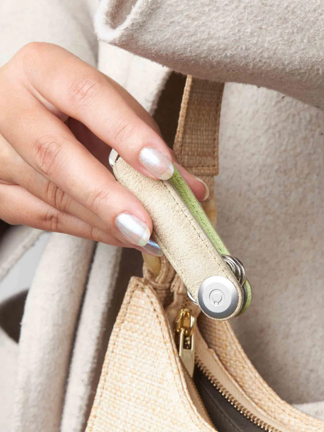 A hand with metallic silver nail polish holds a beige and green multitool near the strap and zipper of a light tan textured bag, next to an Orbitkeys Orbitkey Star Wars Grogu Key Organizer keychain.
