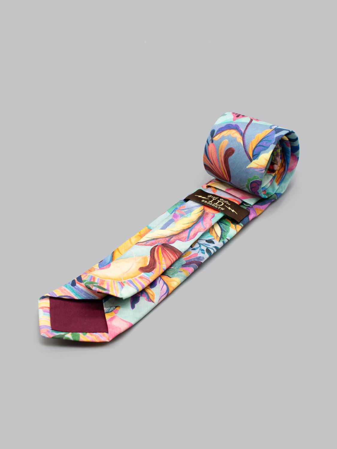 The Liberty of London Tana Lawn™ Una Tie by ID Menswear is a handmade, colorful floral necktie with yellow, pink, and orange flowers on light blue, shown rolled up on a plain gray surface.