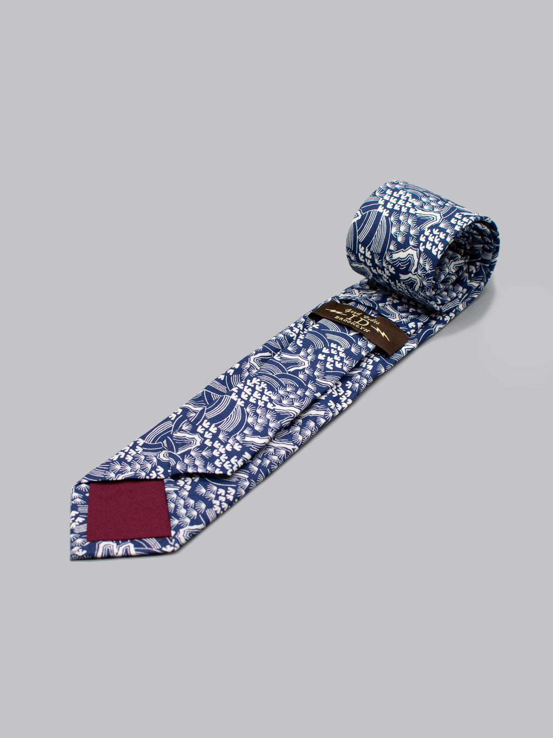 A rolled-up Liberty of London Tana Lawn™ Hillside tie by ID Menswear, blue with a white floral pattern and a dark red tip patch, handmade in Brooklyn from soft cotton, displayed on a plain gray background.