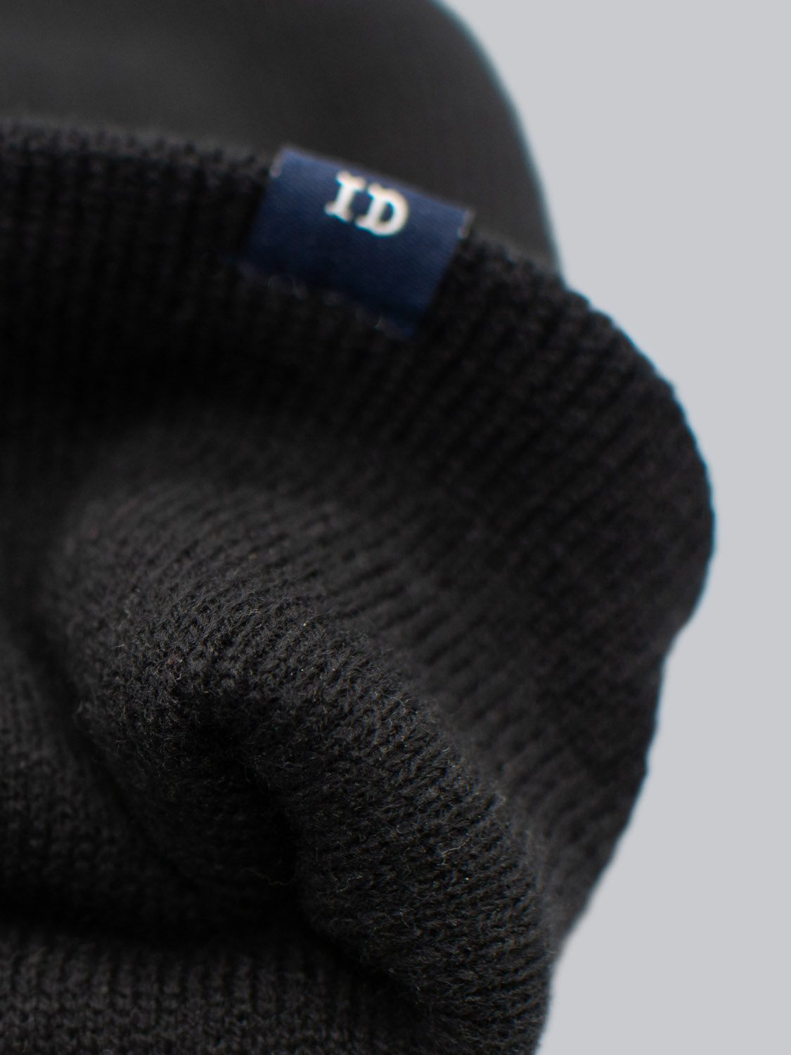 Close-up of the ID Black Beanie by ID Menswear, made in Brooklyn with 100% acrylic fabric. It features a folded edge and a small blue ID tag sewn on. Background: plain light gray.