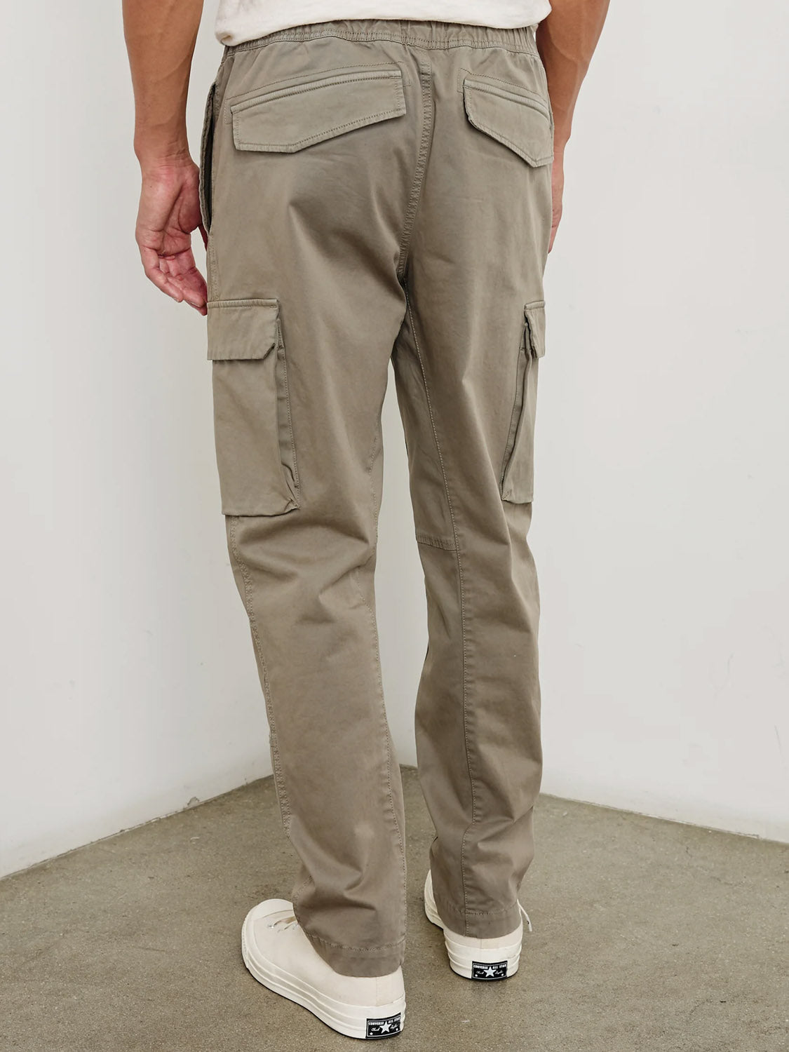 A person stands indoors on a gray floor, facing away from the camera, wearing Rails Jaiden Cargo Pants in light brown with flap pockets and white sneakers.