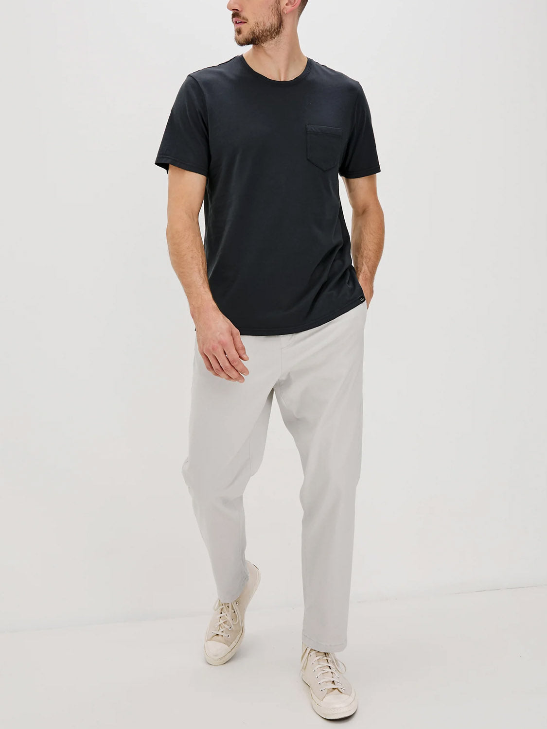 A man stands against a plain white background, wearing the Rails Johnny T-Shirt—a soft cotton jersey tee with a chest pocket—paired with light gray pants and white sneakers. He has one hand in his pocket and looks to the side.
