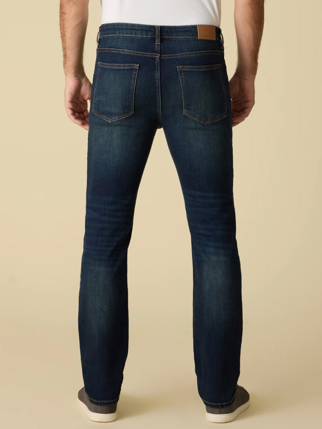 Wearing Flag and Anthem's Jonesville Cooling Jean-Nashville Straight in dark blue, a white shirt, and gray slip-on shoes, a person stands with their back to the camera against a plain beige background.