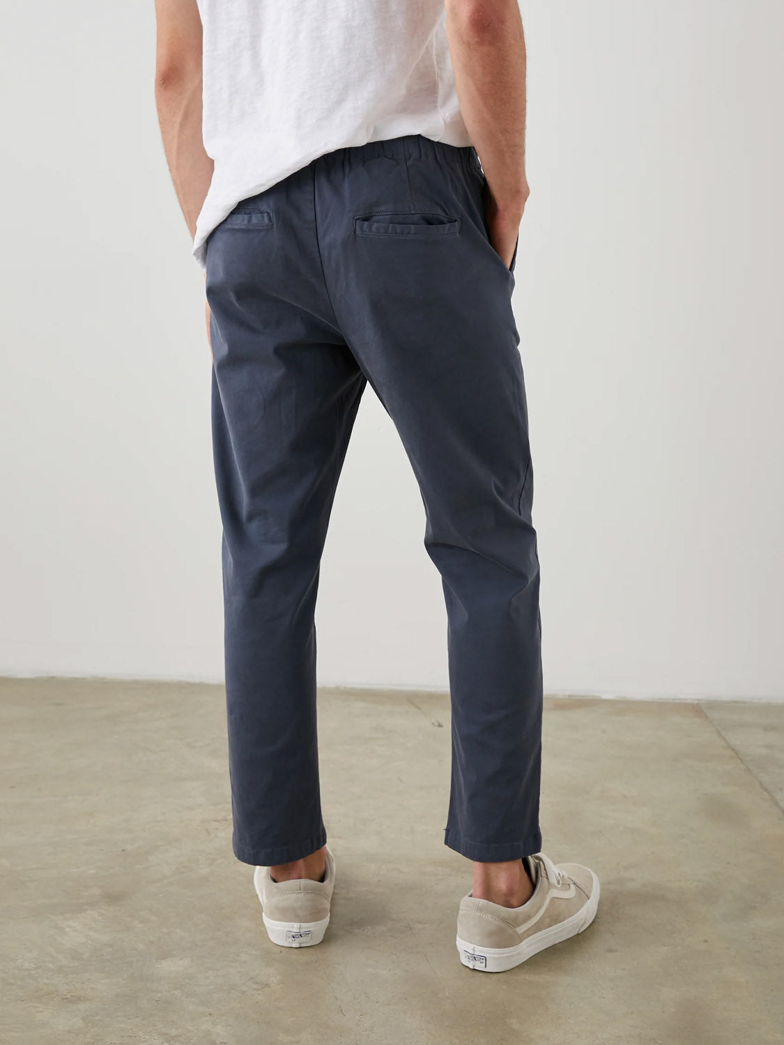 A person stands with their back to the camera, wearing a white T-shirt, Rails Julian Pants in a vintage look, and beige sneakers. Their hands rest in the Rails pants pockets against a plain white wall and concrete floor backdrop.