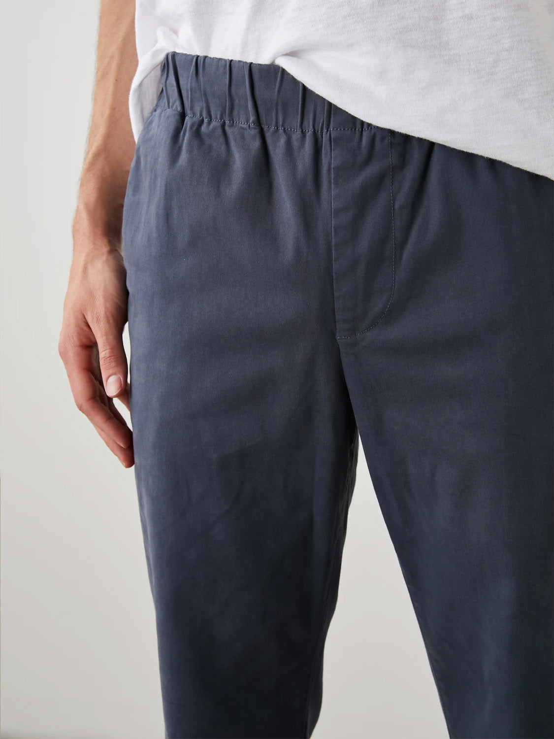 The image shows a person wearing the Rails Julian Pant in dark gray with a vintage look and an elastic waistband, paired with a white shirt. Their left arm hangs by their side.