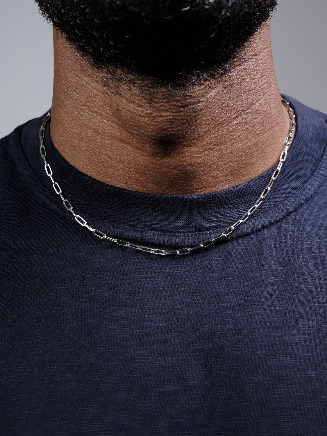 Close-up of a person wearing the Kai 100 Paper Clip Chain Necklace by ID Menswear with a dark navy blue t-shirt, showing the lower half of their face and neck.