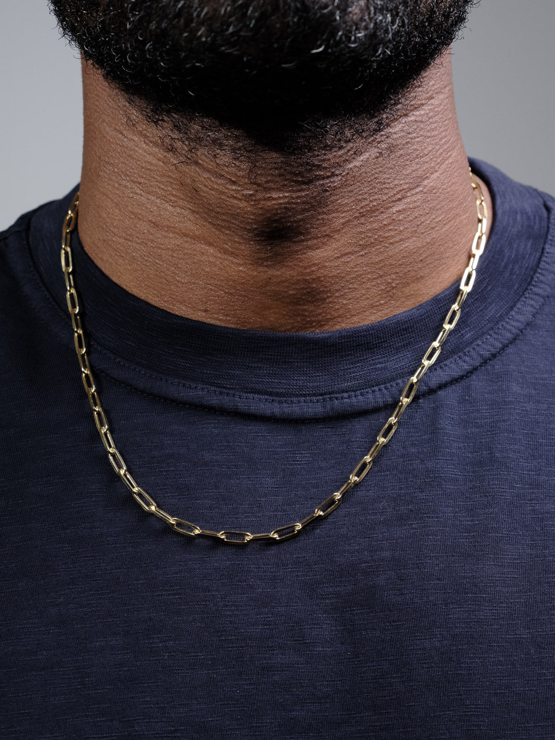 A close-up of a person wearing the Kai 120 Paper Clip Chain Necklace by ID Menswear—a gold plated chain with elongated links—over a navy blue t-shirt. The image focuses on the neck and necklace, with part of the lower face and beard visible.